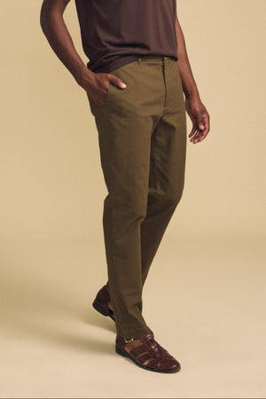 Clad in the LUIS CHINO by Velvet by Graham & Spencer, this individual pairs olive green trousers with a dark brown shirt and brown sandals, standing with one hand in their pocket against a beige background.