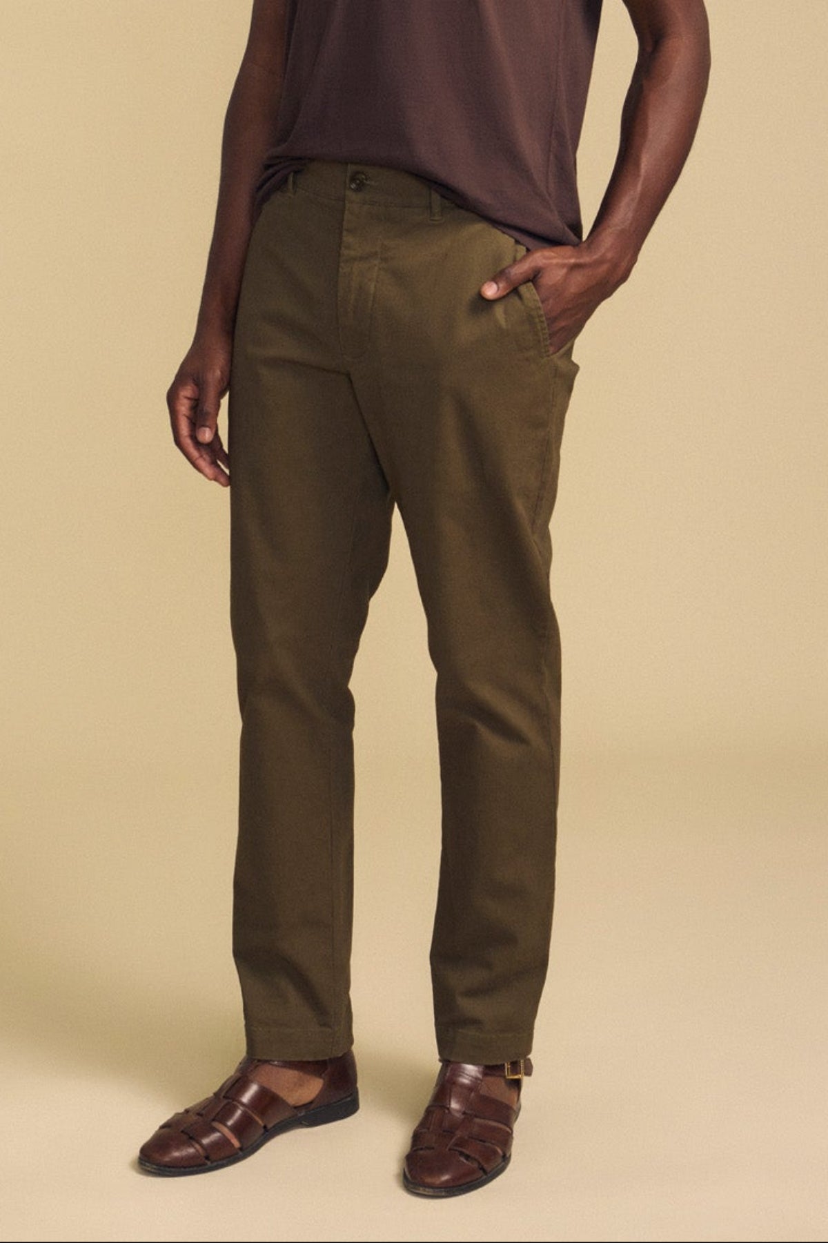 A person wearing the LUIS CHINO trousers by Velvet by Graham & Spencer, paired with a brown short-sleeve shirt and brown sandals, stands with one hand in their pocket against a beige background.