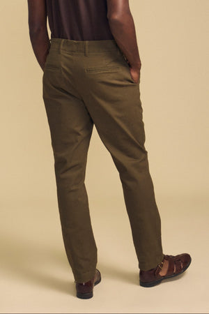 A person stands with hands in pockets, wearing the LUIS CHINO pants in olive green from Velvet by Graham & Spencer and brown sandals, facing away from the camera against a beige background.