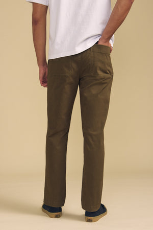 A person stands with their back to the camera, wearing the Velvet by Graham & Spencer CONROY CHINO in brown, paired with a white t-shirt and black shoes, set against a beige background.