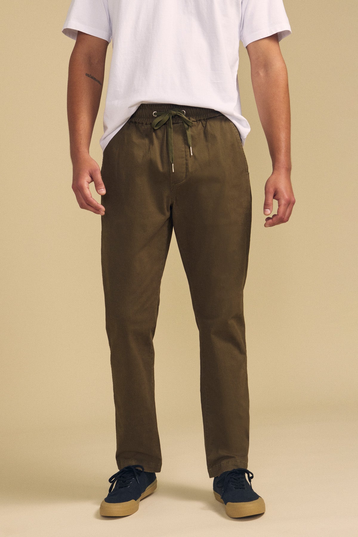 A person wearing a white T-shirt, Velvet by Graham & Spencer CONROY CHINO pants in olive green with a drawstring waist, and dark sneakers is standing against a plain beige background.-41781714813121
