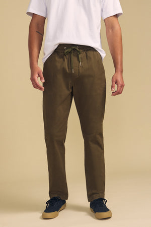 A person wearing a white T-shirt, Velvet by Graham & Spencer CONROY CHINO pants in olive green with a drawstring waist, and dark sneakers is standing against a plain beige background.