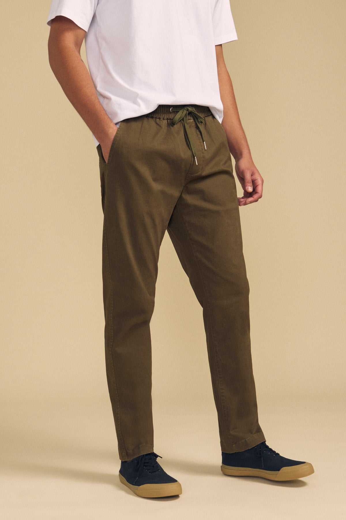 A person wearing a white T-shirt, Velvet by Graham & Spencer’s CONROY CHINO in olive green with a drawstring waist, and black sneakers stands with hands in pockets against a beige background.