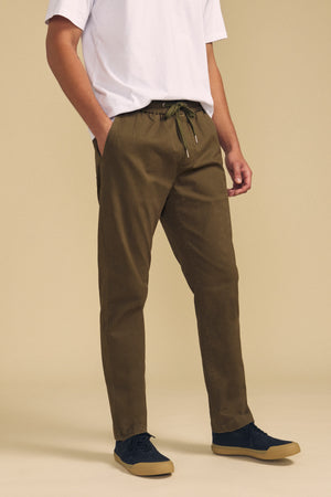 A person wearing a white T-shirt, Velvet by Graham & Spencer’s CONROY CHINO in olive green with a drawstring waist, and black sneakers stands with hands in pockets against a beige background.