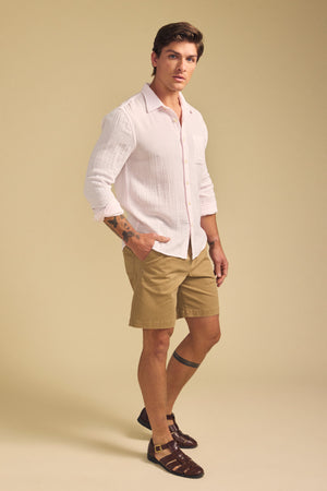 Man standing in a studio wearing the APOLLO SHIRT by Velvet by Graham & Spencer—light pink cotton gauze—paired with tan shorts and brown sandals; neutral background.