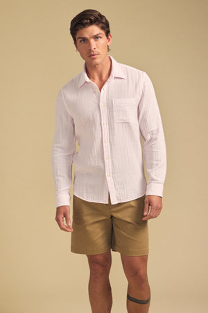 A man stands against a beige background wearing the APOLLO SHIRT in light pink by Velvet by Graham & Spencer, paired with khaki shorts.