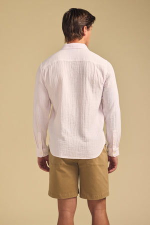 A man stands facing away, wearing the Velvet by Graham & Spencer APOLLO SHIRT—a relaxed cotton gauze long-sleeve—paired with khaki shorts against a plain beige background.