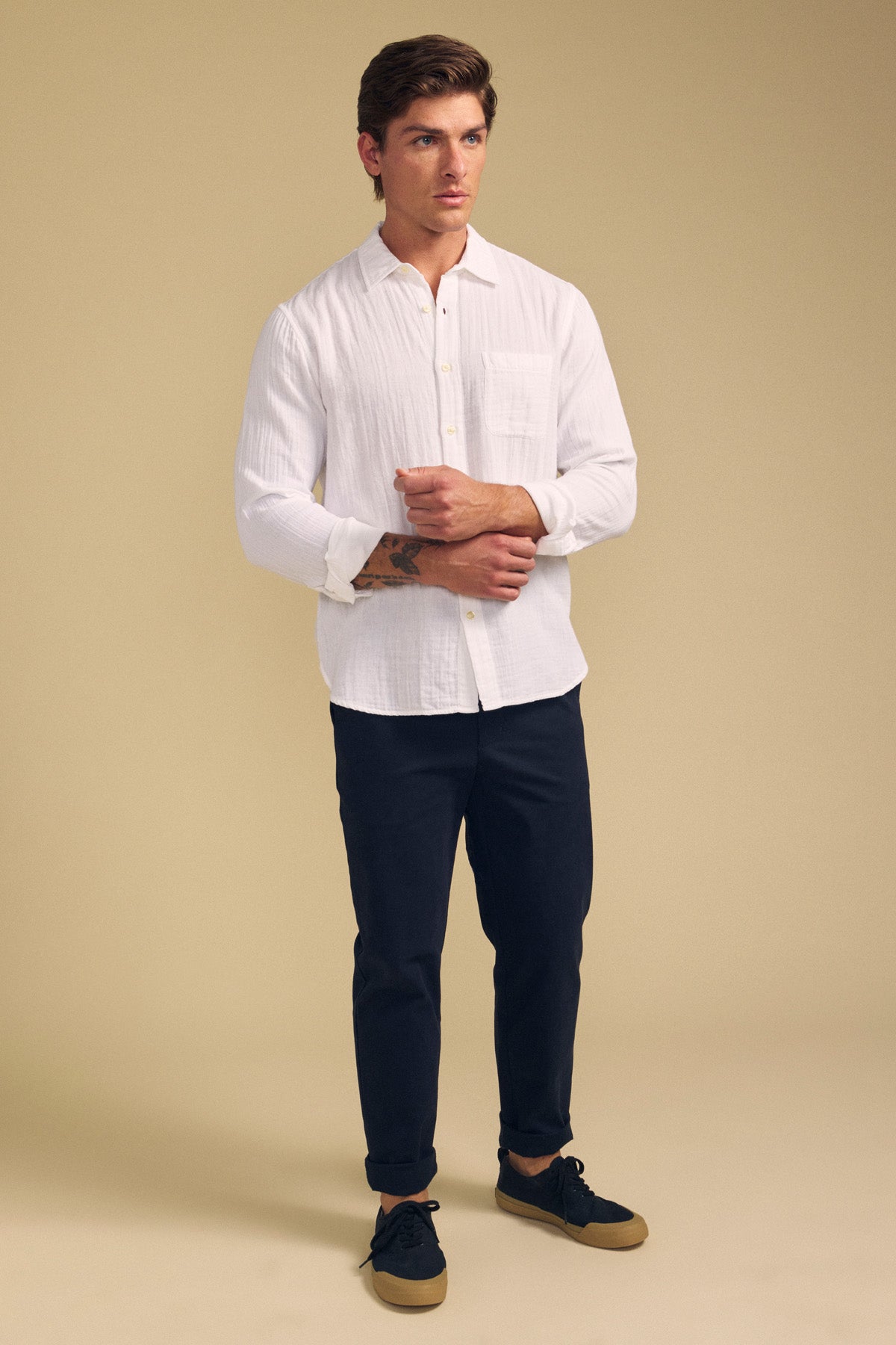 A man stands against a beige background wearing the APOLLO SHIRT by Velvet by Graham & Spencer, paired with dark pants and black sneakers with brown soles.