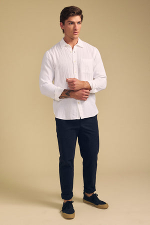 A man stands against a beige background wearing the APOLLO SHIRT by Velvet by Graham & Spencer, paired with dark pants and black sneakers with brown soles.