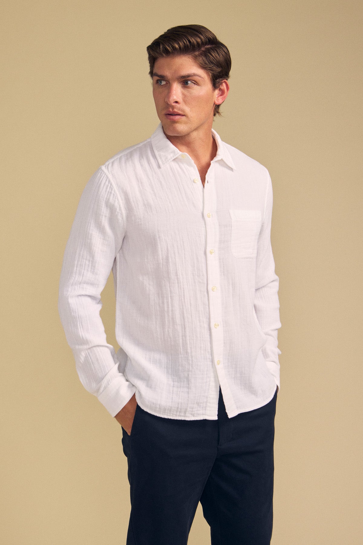 A person wears the APOLLO SHIRT by Velvet by Graham & Spencer in white, paired with dark pants, standing with hands in pockets and looking to the side against a plain beige background.