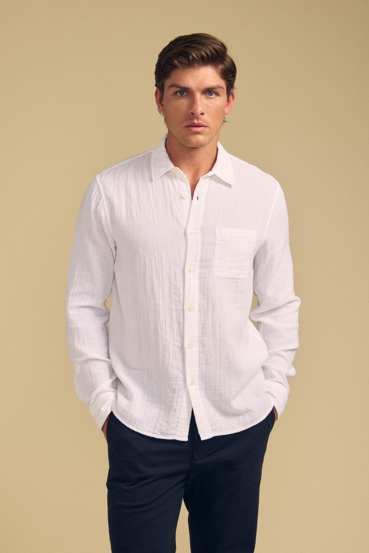A man wears the APOLLO SHIRT by Velvet by Graham & Spencer, a relaxed cotton gauze style with a chest pocket, paired with dark pants against a plain beige background.-41701729173697