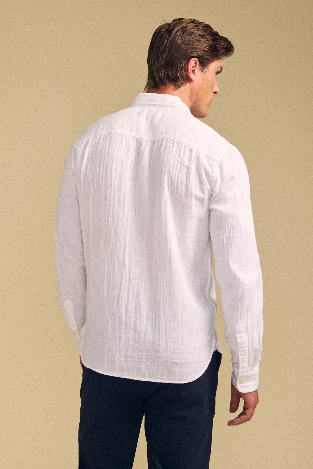 Man stands with back to camera in a long-sleeve white APOLLO SHIRT by Velvet by Graham & Spencer and dark pants, set against a plain beige background.-41701729009857