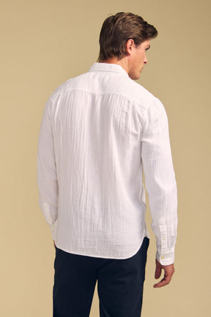 Man stands with back to camera in a long-sleeve white APOLLO SHIRT by Velvet by Graham & Spencer and dark pants, set against a plain beige background.