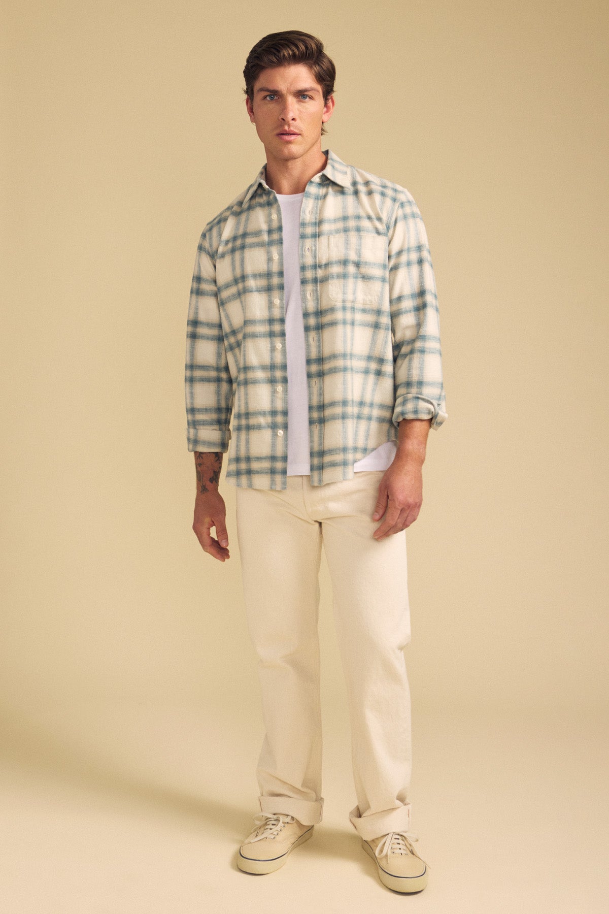 A man wears the COLE SHIRT by Velvet by Graham & Spencer, a light plaid button-up over a white tee, paired with cream pants and beige sneakers, standing against a plain beige background.