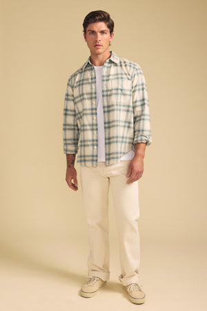 A man wears the COLE SHIRT by Velvet by Graham & Spencer, a light plaid button-up over a white tee, paired with cream pants and beige sneakers, standing against a plain beige background.