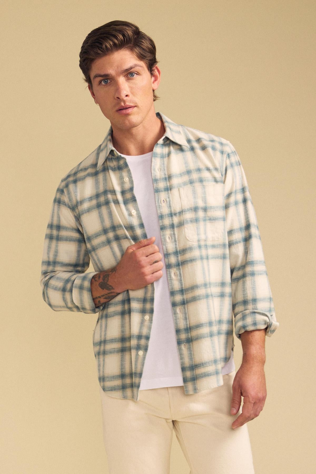 A man stands against a plain beige background wearing the COLE SHIRT by Velvet by Graham & Spencer over a white T-shirt and light pants.-41701909692609