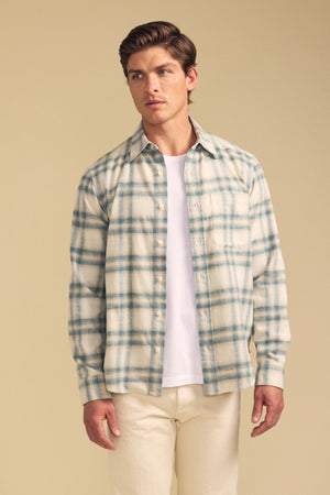 Man wears the COLE SHIRT by Velvet by Graham & Spencer, a cream and blue plaid button-up, layered over a white tee with light pants, standing against a plain beige background.