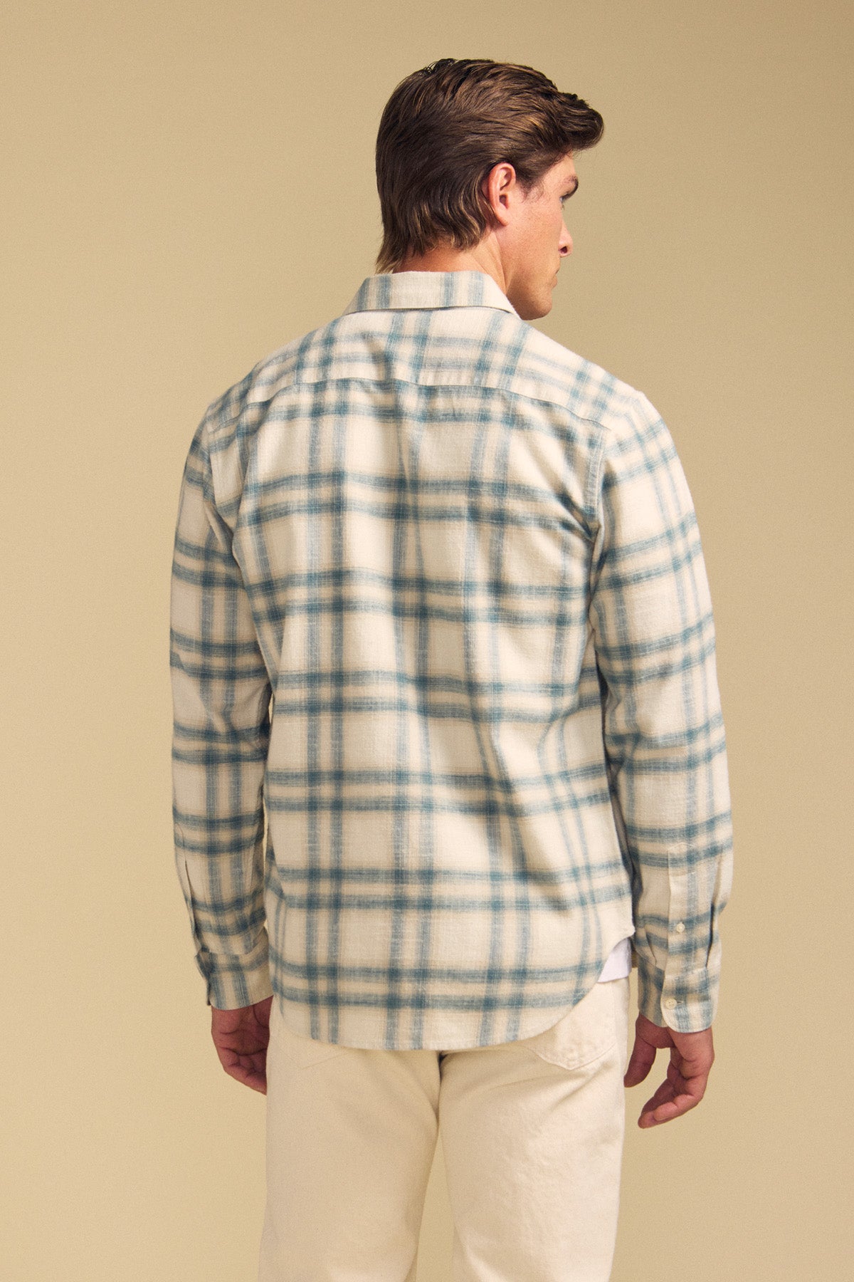 A man faces away, wearing the COLE SHIRT by Velvet by Graham & Spencer—a cream and blue plaid long-sleeve shirt—paired with light pants, set against a plain beige background.-41701907038401