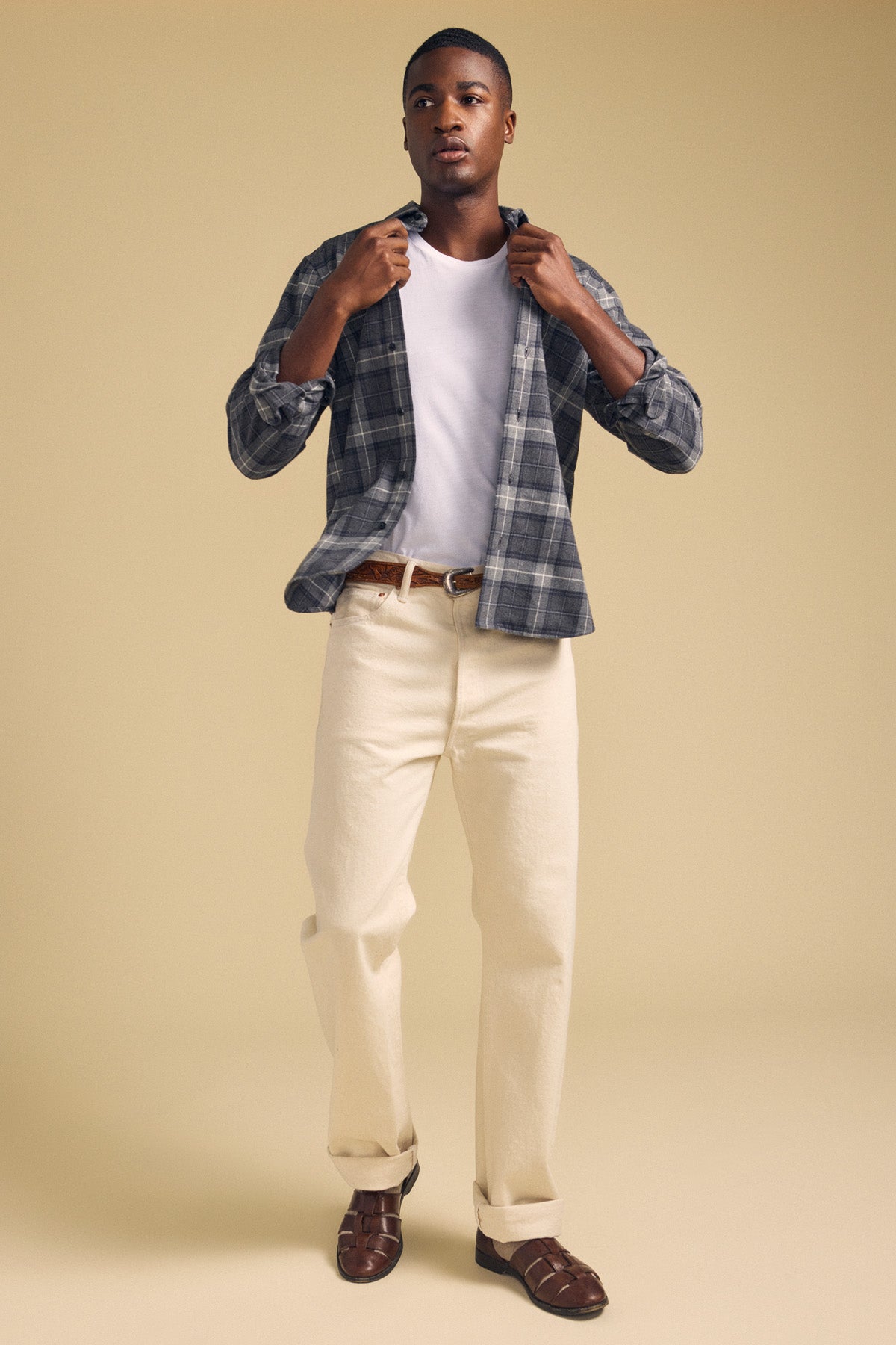 A man stands against a beige background wearing the COLE SHIRT by Velvet by Graham & Spencer—a gray plaid flannel shirt layered over a white t-shirt, paired with cream-colored pants, a brown belt, and brown sandals.