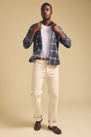 A man stands against a beige background wearing the COLE SHIRT by Velvet by Graham & Spencer—a gray plaid flannel shirt layered over a white t-shirt, paired with cream-colored pants, a brown belt, and brown sandals.
