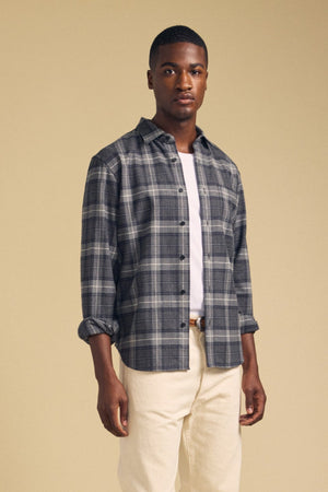 A man stands facing forward wearing the COLE SHIRT by Velvet by Graham & Spencer—a gray plaid flannel men’s shirt—layered over a white T-shirt and beige pants, with a neutral background.