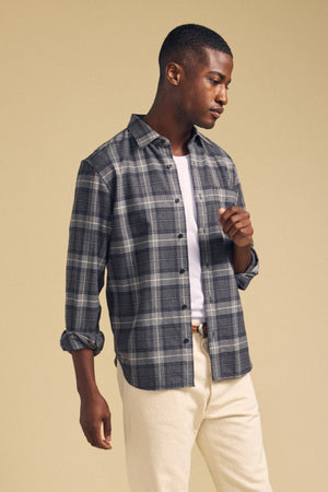 A man stands against a plain beige background wearing the COLE SHIRT by Velvet by Graham & Spencer—a gray plaid flannel men’s button-up crafted from soft brushed cotton—styled over a white t-shirt with rolled sleeves and light-colored pants.