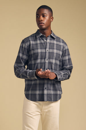 A man stands against a beige background, wearing the COLE SHIRT—a gray plaid flannel button-up by Velvet by Graham & Spencer—and light-colored pants, his hands loosely clasped.