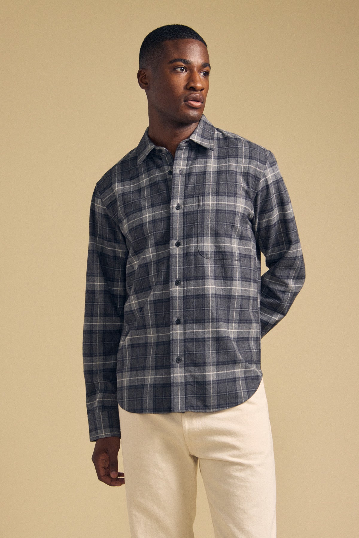 A man dressed in the Velvet by Graham & Spencer COLE SHIRT, a gray plaid flannel button-up, and light-colored pants stands against a plain beige background, looking to his left.