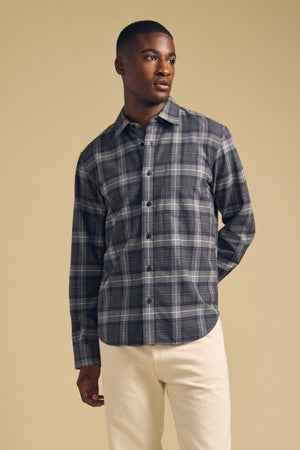 A man dressed in the Velvet by Graham & Spencer COLE SHIRT, a gray plaid flannel button-up, and light-colored pants stands against a plain beige background, looking to his left.