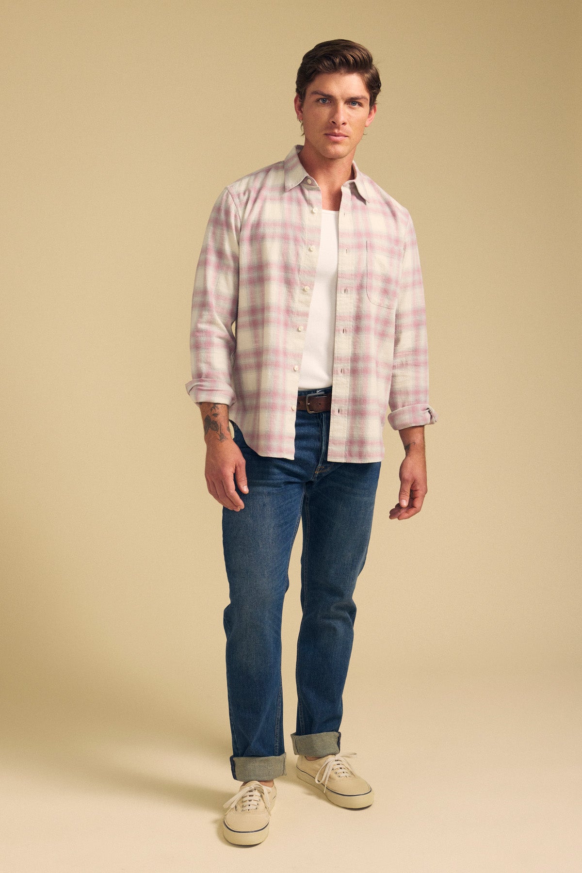 A man stands against a plain beige background wearing the COLE SHIRT by Velvet by Graham & Spencer over a white t-shirt, blue jeans with rolled cuffs, and beige sneakers.
