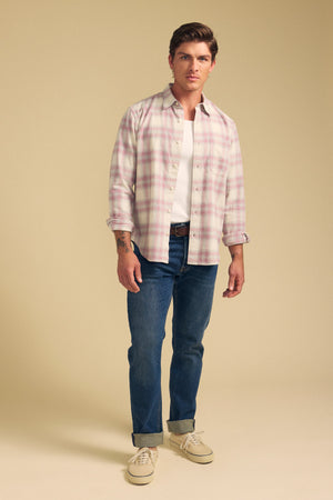 A man stands against a plain beige background wearing the COLE SHIRT by Velvet by Graham & Spencer over a white t-shirt, blue jeans with rolled cuffs, and beige sneakers.