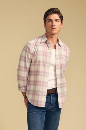 A man stands against a beige background wearing the COLE SHIRT from Velvet by Graham & Spencer—a light pink and white plaid shirt—layered over a white t-shirt and blue jeans.