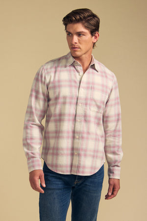 A man models the COLE SHIRT by Velvet by Graham & Spencer—a light pink and white plaid button-up—paired with blue jeans, standing against a plain beige background.