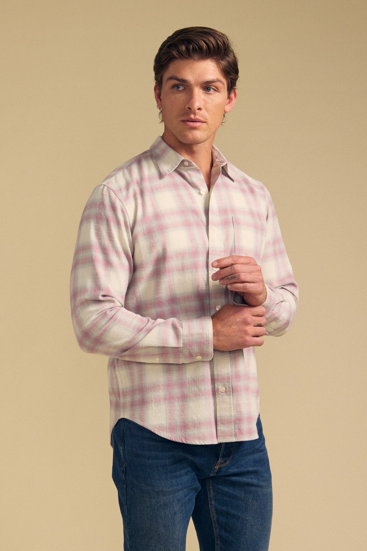 A man wearing the COLE SHIRT by Velvet by Graham & Spencer—a light pink and white plaid button-up—paired with blue jeans stands against a plain beige background.