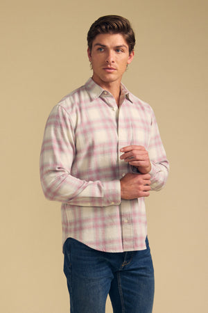 A man wearing the COLE SHIRT by Velvet by Graham & Spencer—a light pink and white plaid button-up—paired with blue jeans stands against a plain beige background.