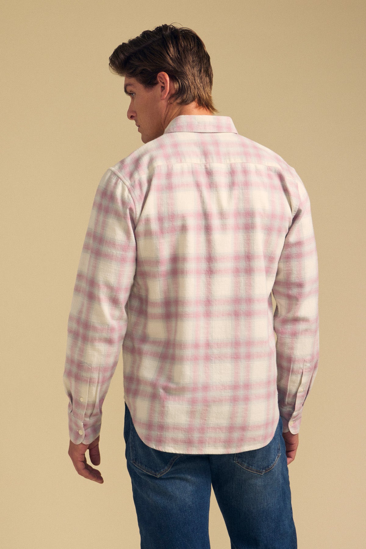 A man stands facing away, wearing the COLE SHIRT by Velvet by Graham & Spencer—a long-sleeve, light plaid shirt with pink stripes—and blue jeans, against a plain beige background.
