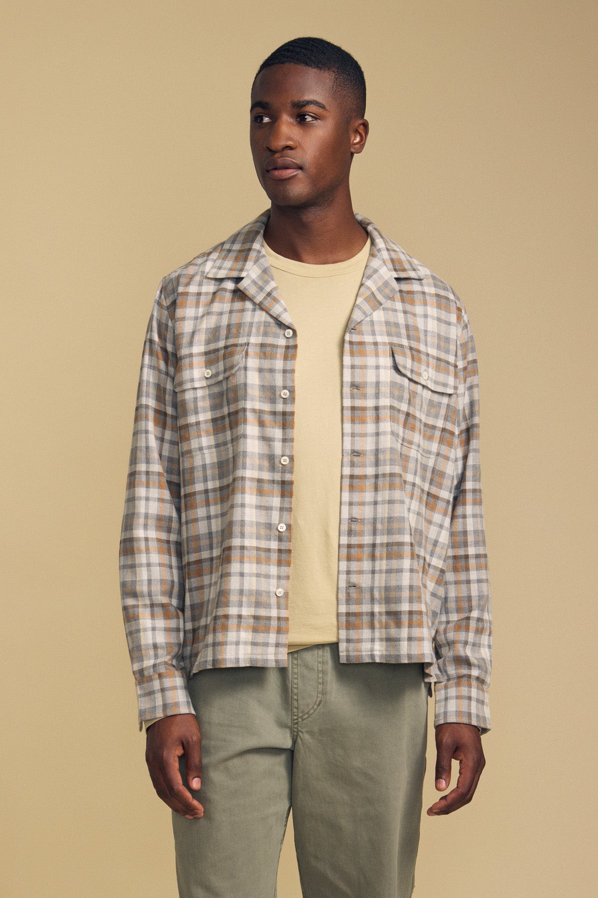 A man stands against a beige background wearing the CUB SHIRT by Velvet by Graham & Spencer—a light plaid button-up—layered over a beige t-shirt and paired with sage green pants.