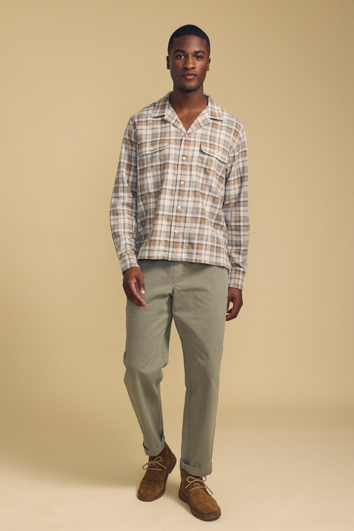 A man stands against a beige background wearing the CUB SHIRT by Velvet by Graham & Spencer, styled with light olive pants and brown suede shoes.