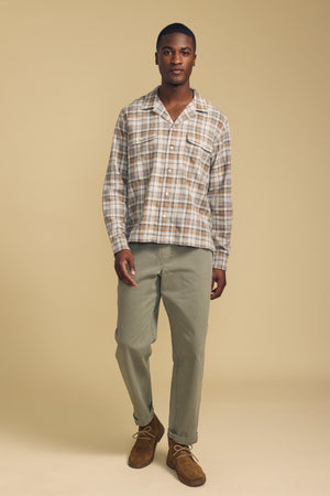 A man stands against a beige background wearing the CUB SHIRT by Velvet by Graham & Spencer, styled with light olive pants and brown suede shoes.