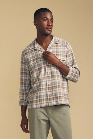 A man wearing the CUB SHIRT by Velvet by Graham & Spencer—a beige and gray plaid shirt with rolled-up sleeves—paired with light green pants stands against a plain tan background.