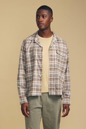 A man stands against a beige background wearing the CUB SHIRT by Velvet by Graham & Spencer—a light plaid button-up—layered over a beige t-shirt and paired with sage green pants.