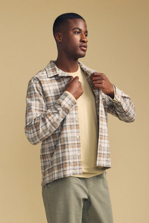 A person wearing the Velvet by Graham & Spencer CUB SHIRT—a light plaid button-down—over a beige t-shirt and green pants stands against a plain beige background, looking to the side.