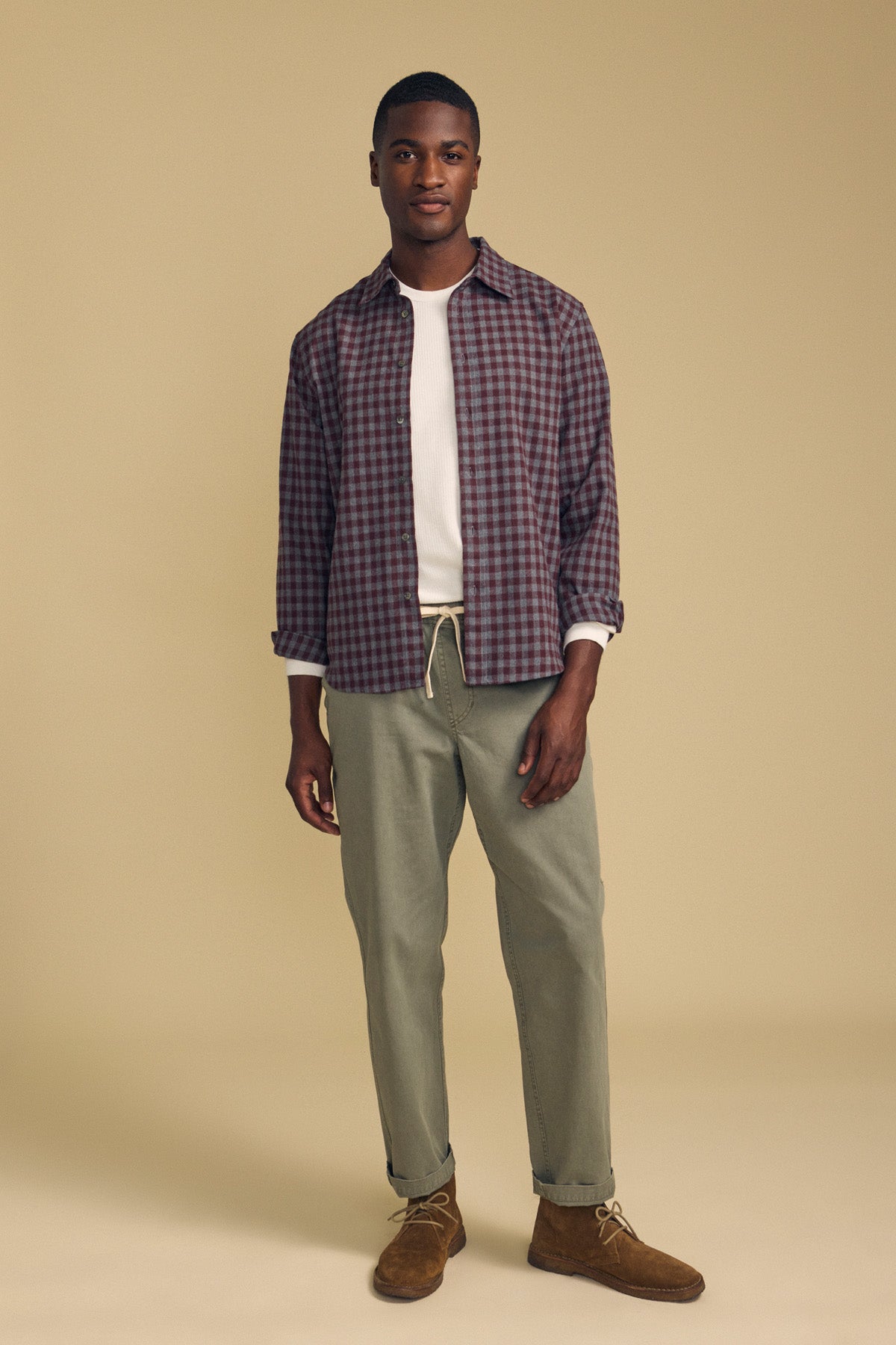 A man wearing a maroon and navy DANNY PLAID BUTTON-UP SHIRT open over a white ribbed shirt, paired with olive green chinos and brown suede chukka boots.