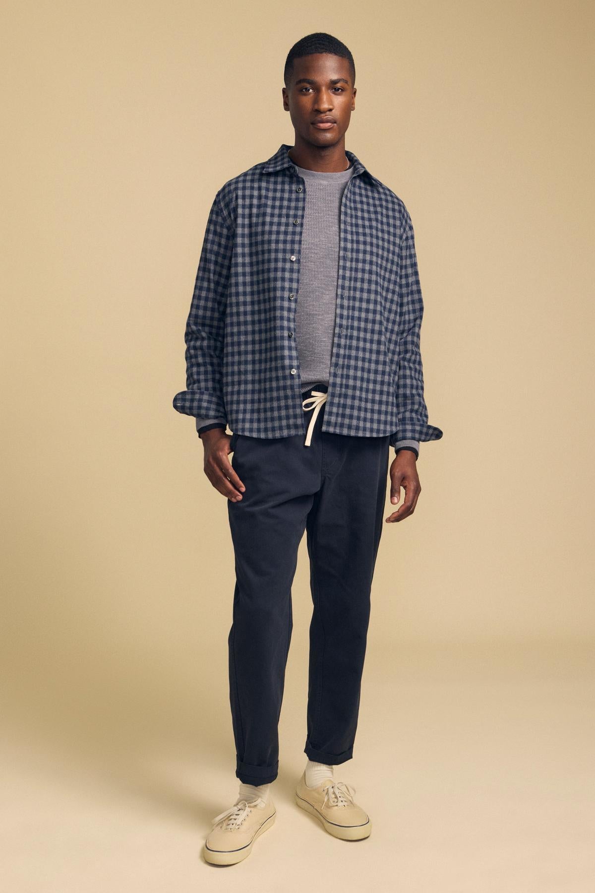 A man wearing a DANNY PLAID BUTTON-UP SHIRT over a grey t-shirt, dark trousers with a drawstring waist, and beige sneakers.