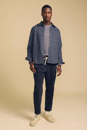 A man wearing a DANNY PLAID BUTTON-UP SHIRT over a grey t-shirt, dark trousers with a drawstring waist, and beige sneakers.