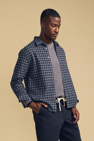 A person wears an unbuttoned blue and gray plaid DANNY PLAID BUTTON-UP SHIRT by Velvet by Graham & Spencer over a gray shirt and dark pants.