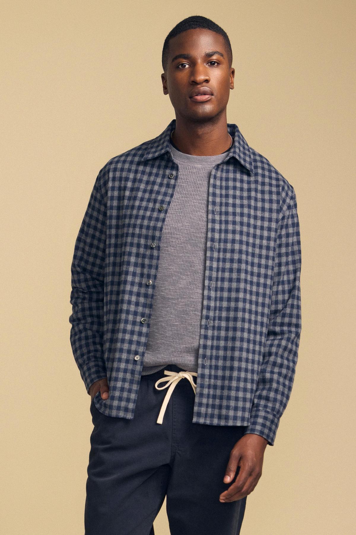 A man wears a blue and grey DANNY PLAID BUTTON-UP SHIRT over a grey t-shirt and dark trousers.-46287908372673