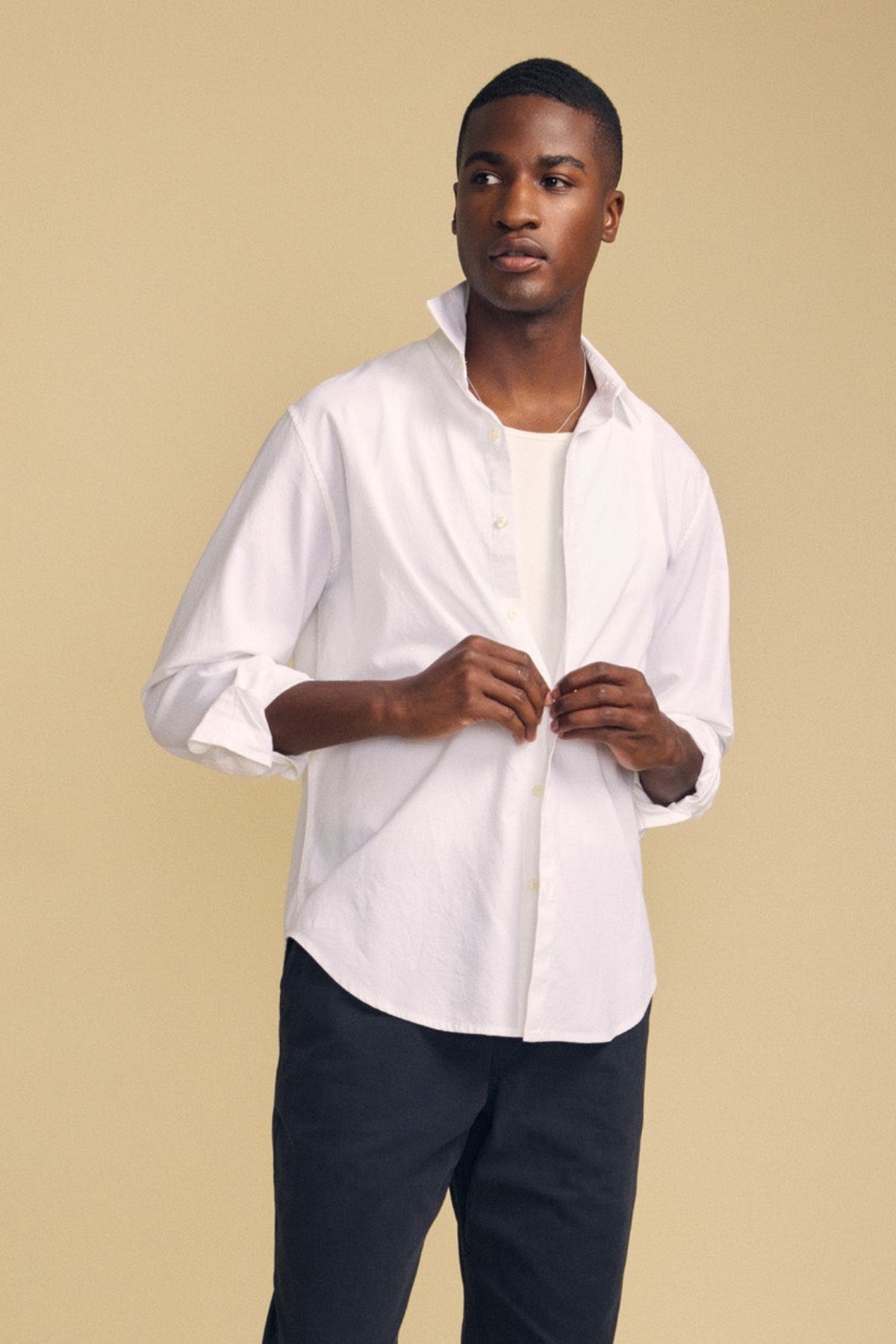 A man stands against a beige background wearing the Velvet by Graham & Spencer RICK SHIRT—a crisp white cotton button-up—layered over a white T-shirt and paired with dark pants, his hands thoughtfully adjusting the shirt.-42092506841281
