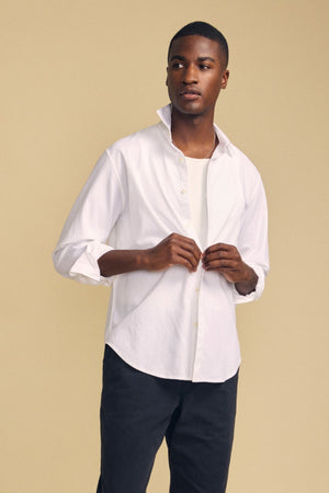 A man stands against a beige background wearing the Velvet by Graham & Spencer RICK SHIRT—a crisp white cotton button-up—layered over a white T-shirt and paired with dark pants, his hands thoughtfully adjusting the shirt.