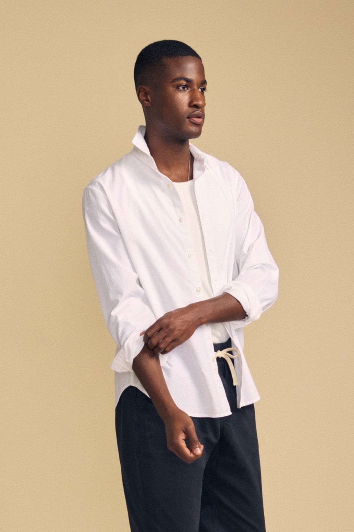 A man stands against a beige background, wearing the RICK SHIRT by Velvet by Graham & Spencer—a crisp white cotton button-up styled over a white top and dark pants, with one sleeve partially rolled up.